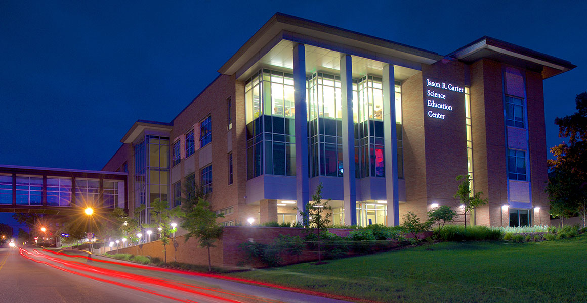 Evening photograph of Metropolitan State University’s Jason R. Carter Science Education Center located on the Saint Paul Campus. Evening sky is dark blue with building light shining bright and remnants of taillights appearing as red streaks from cars driving by on East Sixth Street, West to East.