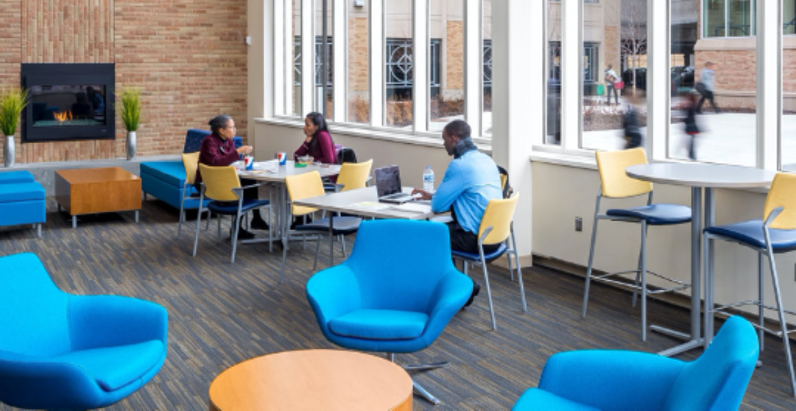 Photograph in Metropolitan State University’s Student Center where two female students are eating their lunches and a male student studying at the next table, with a gas fireplace within a brick wall burning a low flame in an otherwise decorated room with bright blue cushioned chairs, yellow backed and dark blue table chairs, and large windows where passersby can be seen walking towards campus and into the parking ramp. 