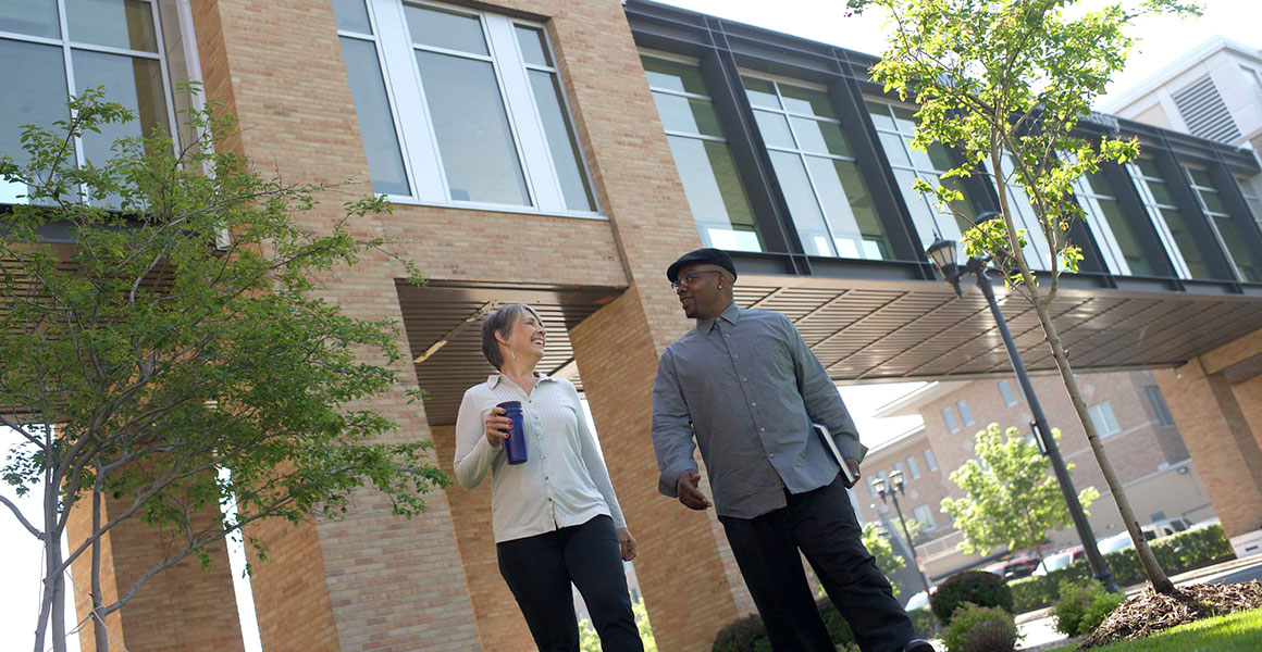 Photograph of an African American male and a Caucasian female smiling, walking and talking on the grounds of Metropolitan State University’s library and learning center with the skyway shown that connects the library to the other building on the Saint Paul campus.