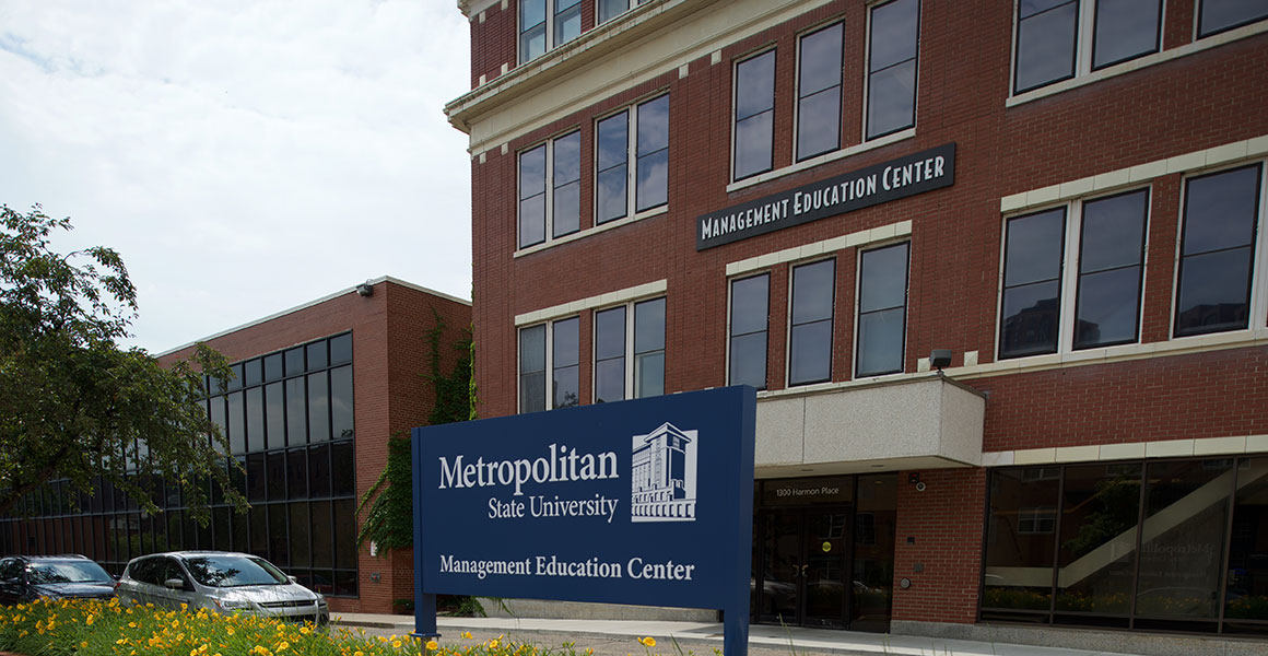 Photograph of Metropolitan State University’s Management Education Center located in Minneapolis, Minnesota; red brick building with white trim, blue sign within an elevated bed of yellow lilies across the street but in front of the building stating, in white lettering, “Management Education Center” with Metropolitan State’s logo shown in white.