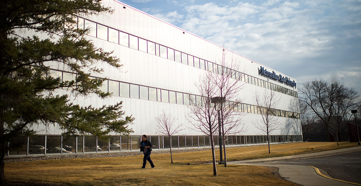 Photograph of the West side of Metropolitan State University’s Midway Center in the very early Spring, with yellow grass and bare trees; a man walks on the sidewalk toward the building from the parking lot.