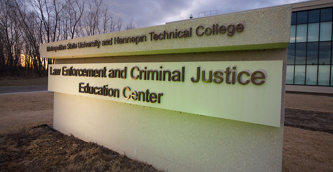 Photograph of the concrete sign outside Metropolitan State University and Hennepin Technical College’s Law Enforcement and Criminal Justice Education Center building taken in the evening, early Spring with yellow grass and bare trees.