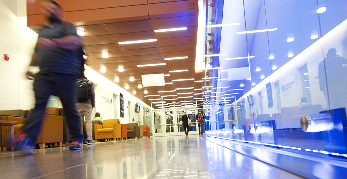 Photograph of students walking in a well-lit corridor at one of Metropolitan State University’s locations; bright lights, reflects, and the blur of a student walking past.