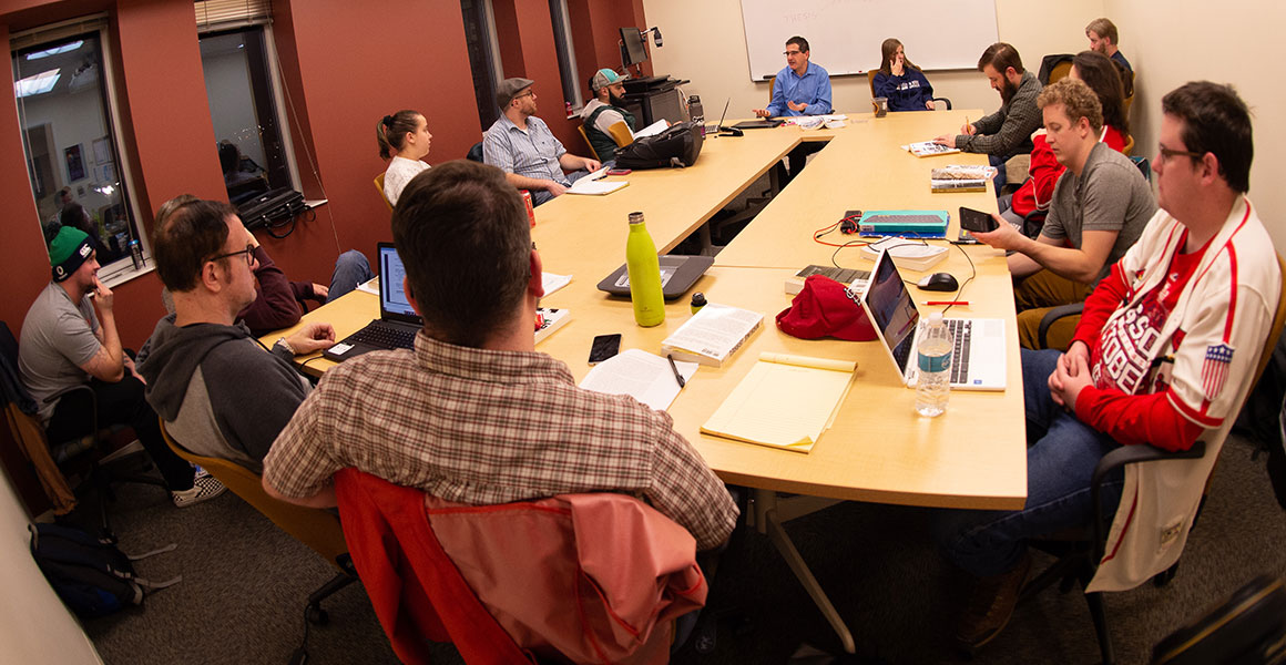Photograph of students gathered around large tables in a smaller meeting room using laptops or taking notes as they enjoy a discussion with the professor in a more non-traditional environment at one of Metropolitan State University’s teaching locations.